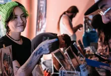 Artist showing artwork to a visitor at a convention table covered with prints and merchandise