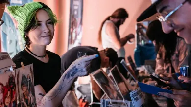 Artist showing artwork to a visitor at a convention table covered with prints and merchandise