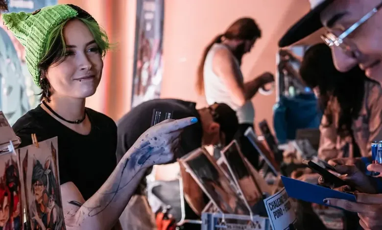 Artist showing artwork to a visitor at a convention table covered with prints and merchandise