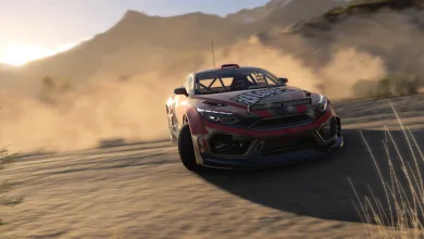 A red-and-black rally car drives across a dusty road with mountains in the background