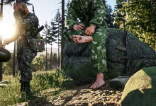 A soldier treats a barefoot teammate beside a rock in a wooded area in Arma Reforger.