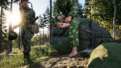 A soldier treats a barefoot teammate beside a rock in a wooded area in Arma Reforger.