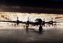 A four-engine bomber sits on a wet hangar floor in front of an illuminated aircraft shelter.