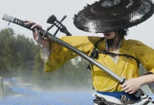 A katana-wielding character in a yellow outfit and straw hat stands in a misty landscape in Ghost of Yōtei.