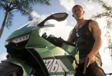 A Grand Theft Auto VI character stands beside a green motorcycle under palm trees while holding a pistol.