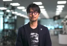 Hideo Kojima wearing glasses and a dark jacket in an illuminated exhibition space