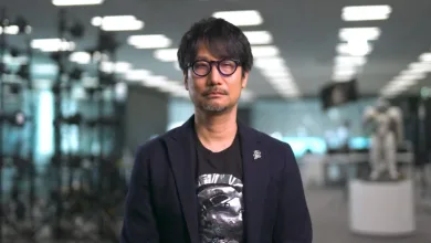 Hideo Kojima wearing glasses and a dark jacket in an illuminated exhibition space