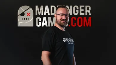 A bearded man wearing a Gray Zone Warfare shirt stands before a MADFINGER GAMING.COM graphic.