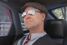 A man wearing a flat cap and sunglasses sits inside a car.
