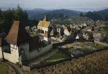 Aerial view of a medieval Manor Lords settlement with a church, fortified buildings, cultivated fields, and wooded hills