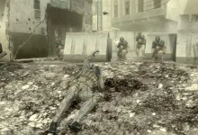 Snake lies prone in a damaged urban street while soldiers approach in Metal Gear Solid 4