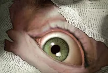 A disturbing green eye peers through strips of gauze in a horror-game scene.
