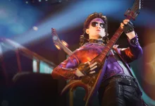 A sunglasses-wearing performer plays an electric guitar onstage in colorful concert lighting.