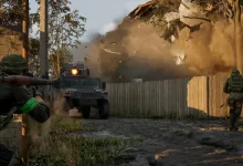 Soldiers take cover near a military vehicle as a structure collapses in a wooded combat area.