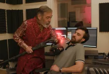 Wes Johnson in a red Sheogorath costume holds a prop beside an audio technician in a recording booth
