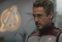 Tony Stark wearing an Avengers uniform with the Avengers emblem visible behind him