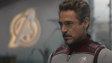 Tony Stark wearing an Avengers uniform with the Avengers emblem visible behind him