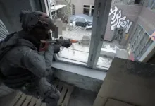 A heavily equipped Bodycam player aims a rifle through a broken window toward a city street.