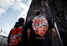 Two people wearing Never Fight Alone Gears of War shirts stand outside a large stone building.