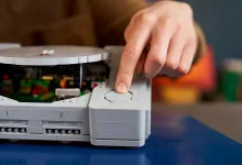 A hand presses a button on a LEGO PlayStation console replica.