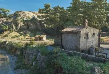 A sunlit countryside landscape with a stone house, stream, rocky hills, and trees in Mafia: The Old Country.