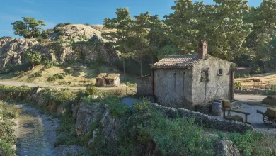 A sunlit countryside landscape with a stone house, stream, rocky hills, and trees in Mafia: The Old Country.