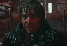 A bloodied, wet-haired man in dark clothing looks ahead in a dim red-lit interior.