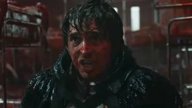 A bloodied, wet-haired man in dark clothing looks ahead in a dim red-lit interior.