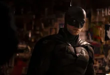 Robert Pattinson in the Batman suit standing in a dimly lit urban setting.