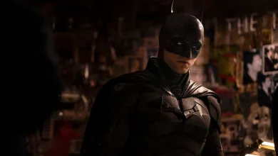 Robert Pattinson in the Batman suit standing in a dimly lit urban setting.