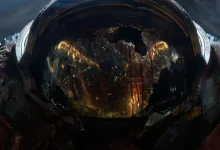 A battered armored helmet with a glowing reflection fills a dark cinematic frame.