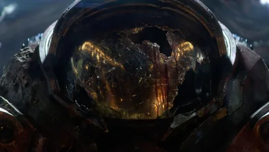 A battered armored helmet with a glowing reflection fills a dark cinematic frame.