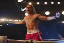 A shirtless fighter in red shorts and boxing gloves raises his arms inside a brightly lit ring.
