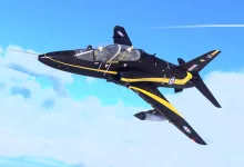 A black-and-yellow Hawk T Mk1A jet flying against a blue sky with clouds.