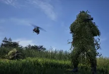 A camouflaged WARDOGS soldier stands in tall grass while another soldier lies nearby and a helicopter flies overhead.