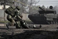 Soldiers from WARDOGS assist a fallen teammate beside a tank in a damaged urban battlefield.