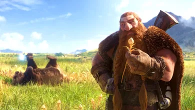 A bearded dwarf holding a small flower stands in a sunlit meadow with mountains in the background.