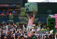 A man raises a large Creeper prop above a crowd at a Minecraft-themed event, beside a Creeper character.