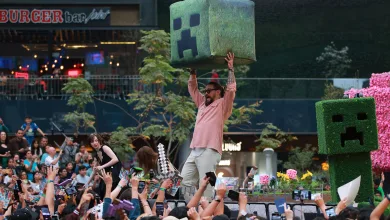A man raises a large Creeper prop above a crowd at a Minecraft-themed event, beside a Creeper character.