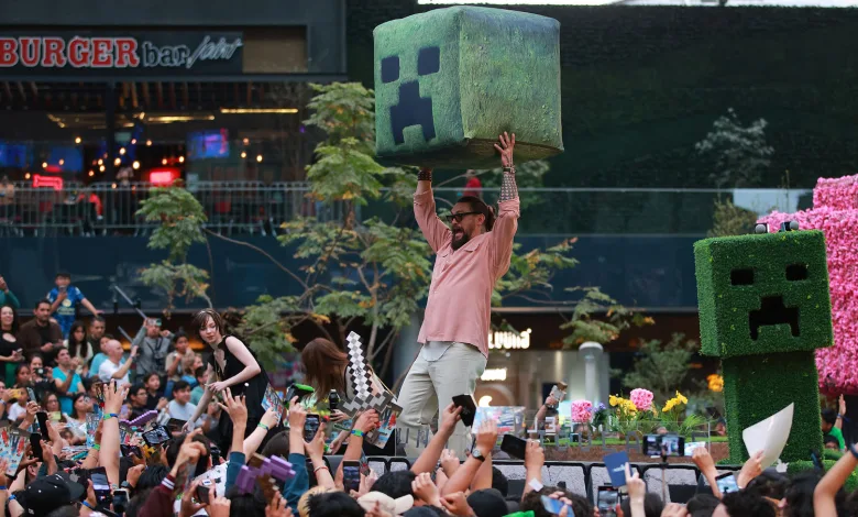 A man raises a large Creeper prop above a crowd at a Minecraft-themed event, beside a Creeper character.