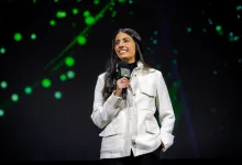 Asha Sharma speaks into an Xbox-branded microphone on a dark stage with green lights behind her.