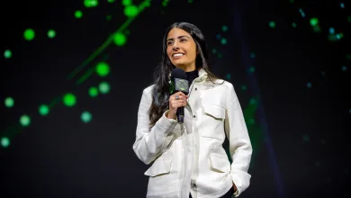 Asha Sharma speaks into an Xbox-branded microphone on a dark stage with green lights behind her.