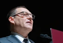Avi Lewis speaks at a microphone against a dark background.
