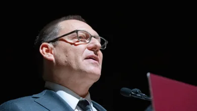 Avi Lewis speaks at a microphone against a dark background.