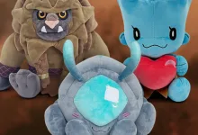 Three Enshrouded plush toys arranged together, including a blue creature holding a red heart