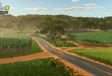A paved road winds through green fields and red soil beneath a forested skyline in Farming Simulator 25.