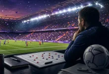 A football manager studies a tactics board while watching a match in a packed stadium.