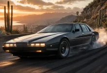 A dark gray Aston Martin Lagonda slides through a smoky drift on a desert road, with a sunset over a cactus-covered valley.