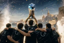 Five esports players in black jerseys celebrate together as one raises a trophy above them, with confetti and a coastal city in the background.