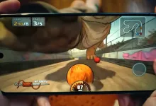 A smartphone held in two hands displays a colorful racing game with a speedometer and track map on screen.